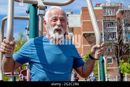 Un uomo anziano esausto che cattura il respiro dopo aver fatto esercizio sulle attrezzature pubbliche della palestra all'aperto. Foto Stock