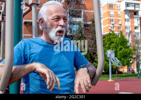 Un uomo anziano esausto che cattura il respiro dopo aver fatto esercizio sulle attrezzature pubbliche della palestra all'aperto. Foto Stock