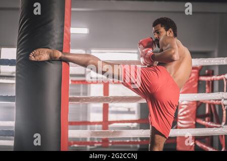 Il giovane kickboxer dalla pelle scura calcia un sandbag con la gamba Foto Stock