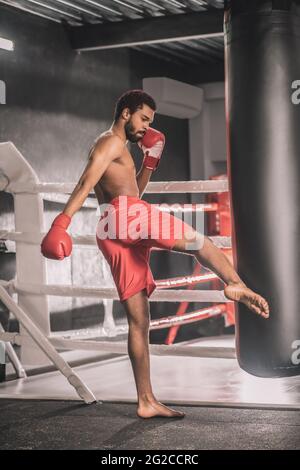 Il giovane kickboxer dalla pelle scura calcia un sandbag con la gamba Foto Stock