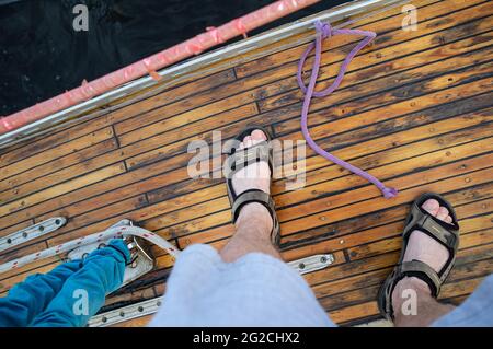 I piedi maschi in sandali si trovano sul ponte di legno di uno yacht sullo sfondo di acque scure. Vista dall'alto. Foto Stock