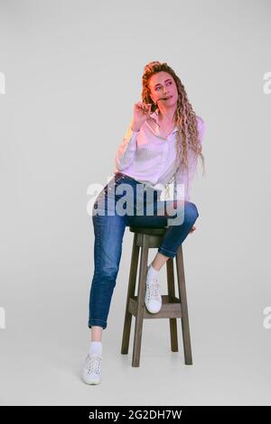 Donna felice in jeans e camicia di cotone con dreadlock si siede sulla sedia sotto le luci colorate Foto Stock