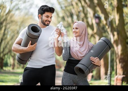 Felice uomo e donna musulmana tostare con una bottiglia di acqua dolce dopo l'allenamento al parco verde. Giovane coppia in activewear che tiene tappetini yoga mentre rinfrescando all'aperto. Foto Stock