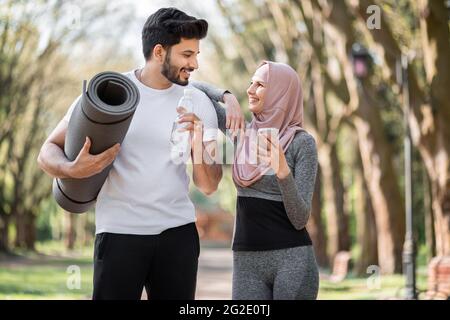 Bel ragazzo arabo con tappetino yoga e bottiglia d'acqua guardando la sua moglie affascinante in hijab che utilizzando il telefono cellulare. Giovane famiglia in activewear in piedi al verde del parco del mattino. Foto Stock