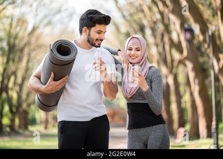 Giovane donna in abbigliamento sportivo e hijab utilizzando smartphone moderno, mentre bell'uomo musulmano bere acqua e tenere tappetino yoga. Una coppia felice che si rilassa dopo un allenamento all'aria aperta. Foto Stock