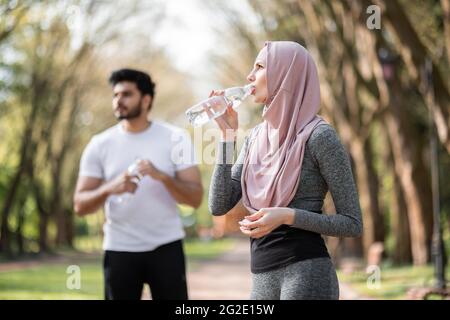 Vista laterale di bella donna in hijab acqua potabile, mentre bel ragazzo arabo in piedi dietro. Giovane coppia in abbigliamento sportivo rilassante dopo l'allenamento all'aria aperta. Foto Stock