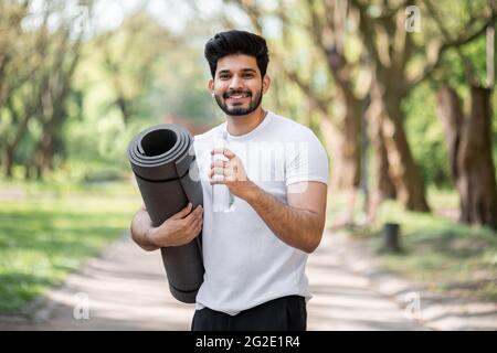 Ritratto di sorridente arabo in abiti sportivi acqua potabile dopo l'allenamento al parco. Giovane ragazzo attivo che posa all'aperto con tappetino yoga in mani. Foto Stock
