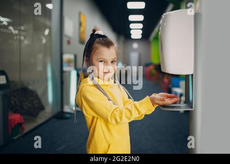 La bambina usa l'igienizzatore automatico con l'erogatore di gel di alcool per le mani in asilo. Disinfettante antisettico per interni, nuovo concetto di pandemia normale di Coronavirus COVID-19. Foto Stock