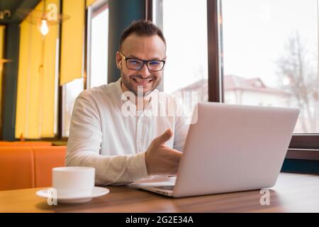 Ritratto di un giovane bel uomo d'affari in una camicia bianca, bicchieri, con la barba si siede a un tavolo in un computer portatile su una tazza di caffè e sorrisi. Foto Stock