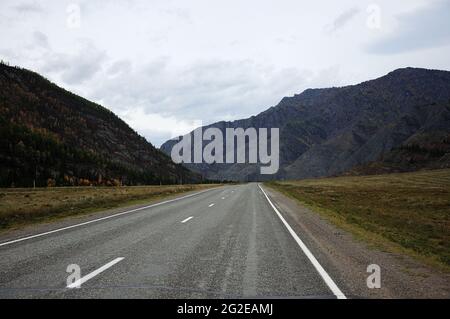 Una strada asfaltata diritta a due corsie con segnaletica attraversa una valle in uno stretto canyon. Chuisky, Altai, Siberia, Russia. Foto Stock