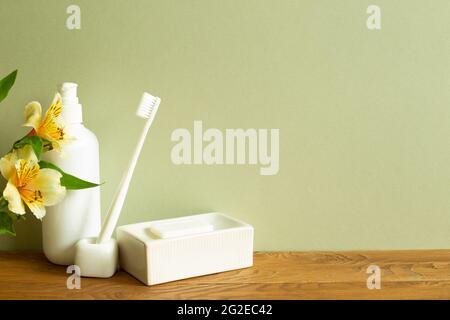 Bottiglia di shampoo, spazzolino da denti, sapone, fiore su tavolo di legno. Sfondo verde parete kaki. Interno del bagno. Cura della pelle e concetto spa Foto Stock