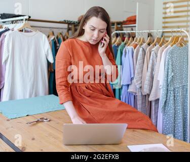 Donna fashion designer che controlla l'ordine online sul computer portatile mentre parla con il cliente sul telefono cellulare nella sua officina Foto Stock