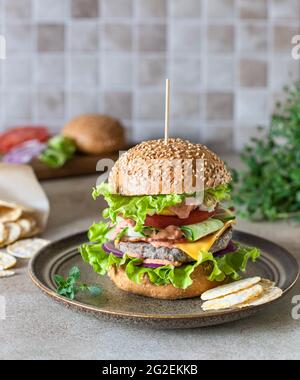 Gustoso hamburger di manzo fresco fatto in casa su piastra di ceramica con ingredienti per la preparazione su sfondo. Fast food e concetto di cibo spazzatura. Messa a fuoco selettiva. Foto Stock