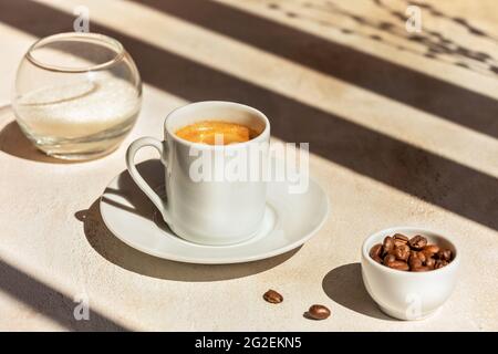 Il sole, la mattina, il caffè è ancora vivo con una tazza di caffè espresso, chicchi di caffè, zucchero e lunghe ombre scure. Caffè forte, arabico e appena preparato in una tazza Foto Stock