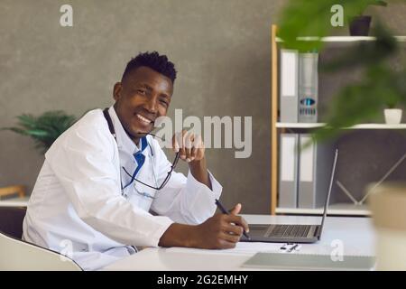 Giovane medico nero seduto alla scrivania dell'ufficio con il computer portatile, guardando la fotocamera e sorridendo Foto Stock