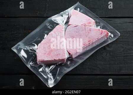 Bistecca di tonno surgelata in un set di confezioni di plastica sottovuoto, su fondo di legno nero Foto Stock