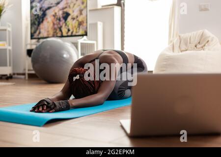 Donna nera che esercita e allunga il suo corpo facendo esercizi di flessibilità seduta su tappetino yoga nel soggiorno domestico che indossa leggings durante il tutorial online. Allenamento e stile di vita sano. Foto Stock