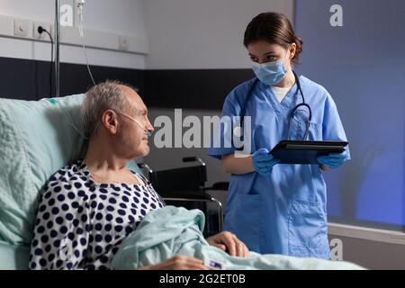 Assistente medico con maschera di protezione controllo trattamento di uomo anziano leggere note su tablet pc. Ammalato anziano che si trova a letto discutendo con l'infermiere circa l'esperienza di diagnosi con la sacca per infusione attaccata. Foto Stock