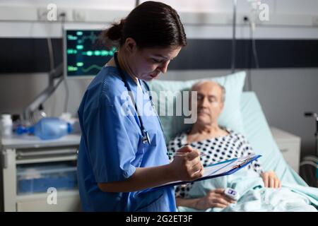 Assistente medico che controlla il trattamento dell'uomo anziano che legge le note sulla clipboard. Ammalato anziano che si trova a letto discutendo con l'infermiere circa l'esperienza di diagnosi con la sacca per infusione attaccata. Foto Stock