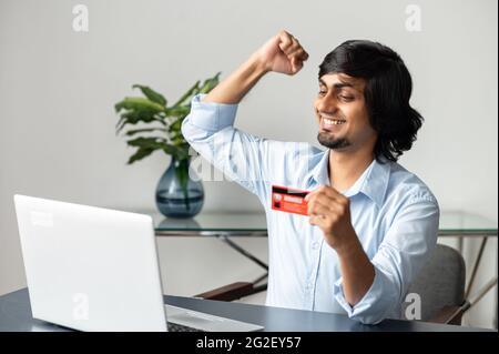 L'imprenditore indiano maschio molto gioioso felice con il pagamento ricevuto sulla sua carta bancaria, tiene la carta e guarda lo schermo del portatile, alzando pugno in gesto di sì. Uomo d'affari orientale shopping online Foto Stock