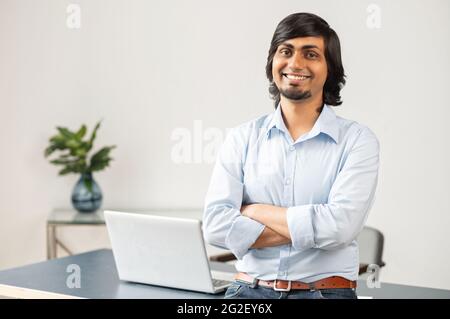 L'imprenditore indiano maschio ambizioso e altamente qualificato si leva in posizione fiduciosa in ufficio moderno, sorridente uomo d'affari orientale in abbigliamento casual guarda la macchina fotografica con le braccia incrociate. Successo e successo Foto Stock