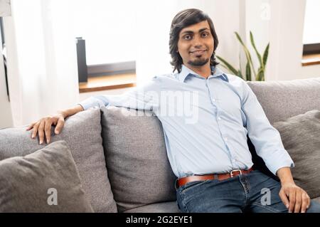 Carismatico uomo indiano dai capelli scuri in elegante abbigliamento casual si siede sul divano e guarda la macchina fotografica. Ritratto di bel ragazzo indù Foto Stock