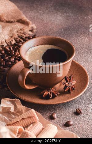 Caffè in una tazza di ceramica marrone con un piattino, accanto ad esso anice, chicchi di caffè e biscotti su uno sfondo di pietra. Foto Stock