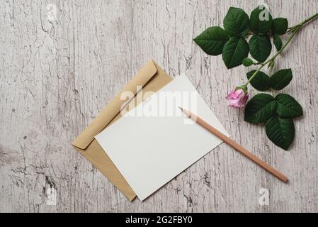 Mockup bianco biglietto d'auguri o invito, busta marrone kraft, matita e rosa fiore su sfondo grigio. Disposizione piatta, vista dall'alto. Spazio di copia Foto Stock