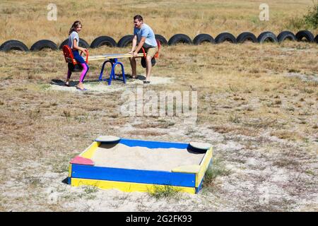 Defocalizzare la bambina che oscilla sul campo da gioco con il giovane uomo, ragazzo, suo fratello maggiore. Zona di campagna. Swing e sandbox blu e rosso brillante. F Foto Stock