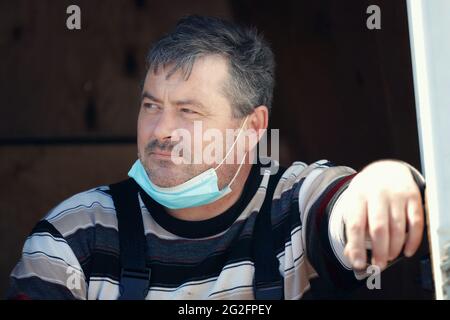 Un uomo bianco di mezza età che indossa una maschera medica guarda via pensieroso. Pesanti pensieri del lavoratore sul futuro durante la pandemia Foto Stock