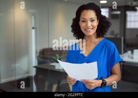 Ritratto di sorridente donna d'affari afroamericana in possesso di documenti in piedi in ufficio Foto Stock