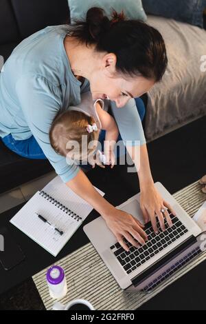 Sorridente madre caucasica che tiene il suo bambino usando il laptop mentre lavora da casa Foto Stock