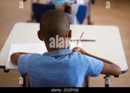 Vista posteriore del ragazzo afroamericano che studia mentre si siede sulla sua scrivania nella classe a scuola Foto Stock