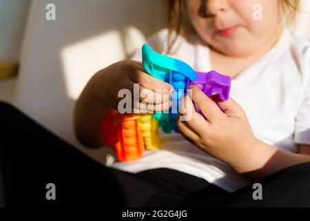 Bambina con giocattolo popit in colori arcobaleno. Giocattolo in silicone a forma di dino per ridurre lo stress. Bolle sensoriali fidget trendy. Bambino che gioca a casa Foto Stock