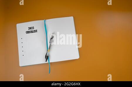 Un libro di note con una penna e una citazione di testo '2022 Checklist' isolato su sfondo marrone. Focus selettivo sul libro delle note. Foto Stock