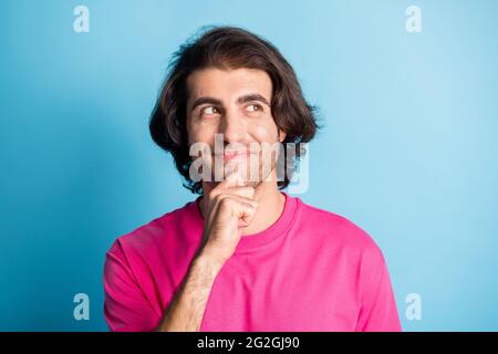 Ritratto di capelli brunet positivi giovane uomo tenere mano mento sguardo vuoto spazio indossare t-shirt rosa isolato su sfondo blu Foto Stock