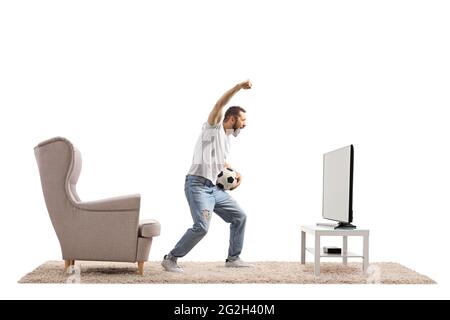 Giovane uomo di fronte a una tv che tiene una palla di calcio e che si acclama con la mano su isolato su sfondo bianco Foto Stock