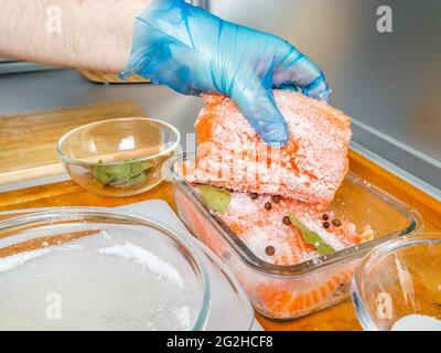 La cuoca in guanti igienici prepara il salmone leggermente salato. Pezzi di pesce cosparsi di sale sono disposti in una ciotola di vetro. Foto Stock