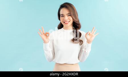 Giovane donna felice in t-shirt bianca, strizzandosi e mostrando i segni giusti, sorridente soddisfatto, raccomanda la pubblicità, approva il prodotto su sfondo blu Foto Stock