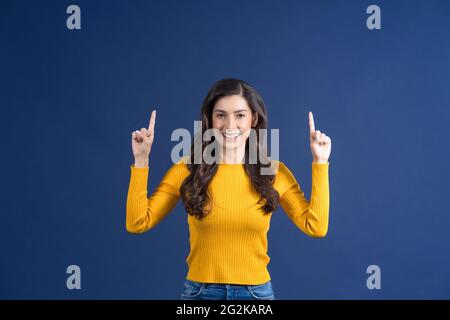 Felice giovane donna asiatica sorridente dito puntando a fare pubblicità con felice emozione su sfondo blu colore, mostrando o presentando il prodotto via vuoto Foto Stock