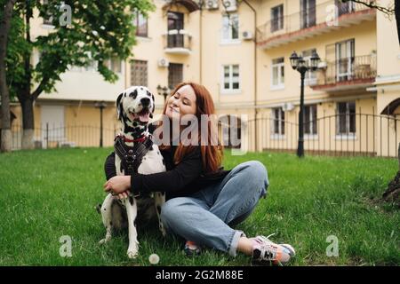 Donna felice che posa e gioca con il suo cane dalmata mentre si siede in erba verde durante una passeggiata urbana della città. Concetto di amicizia, amore e cura Foto Stock