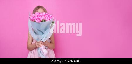 Una bambina in abito rosa con bouquet di rose, nascose dietro il flowers.The ragazza inala la fragranza di fiori su bandiera rosa backgraund Foto Stock