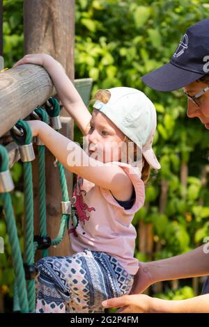 Una madre che aiuta il suo bambino di 3 anni a salire le corde su un telaio di arrampicata. Tema: Relazione madre-bambino, guida parentale, insegnamento, apprendimento Foto Stock