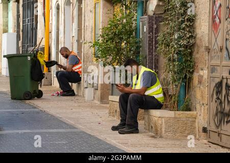 Gerusalemme, Israele - 3 giugno 2021: Due pulitori di strada della città che prendono una pausa dal lavoro per usare i loro telefoni cellulari. Foto Stock