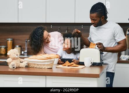 Famiglia afroamericana con bambina sono preparati panini in cucina insieme, tostando il pane e trascorrendo il tempo divertente insieme Foto Stock