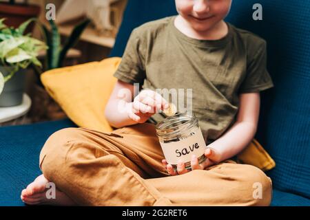 Primo piano delle mani del bambino piccolo ragazzo che afferra e mette le monete della pila in vaso di vetro con l'etichetta di salvataggio. Donazione, risparmio di denaro, beneficenza, famiglia Foto Stock