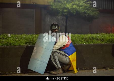 Un dimostratore dalla prima linea che usa uno scudo di mano si erge con una bandiera colombiana mentre le persone si scontrano con la polizia maledetta della Colombia (ESMAD) sulla strada che collega Cartago, Valle del Cauca con la città di Pereira, Risaralda, Colombia 11 giugno 2021. Foto Stock