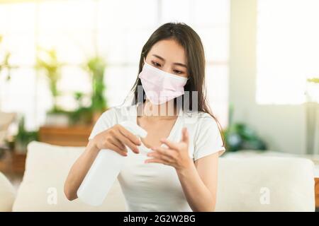 Giovane donna che lavora a casa, indossando una maschera seduta sul divano, spruzzando le mani con alcol per disinfettare Foto Stock
