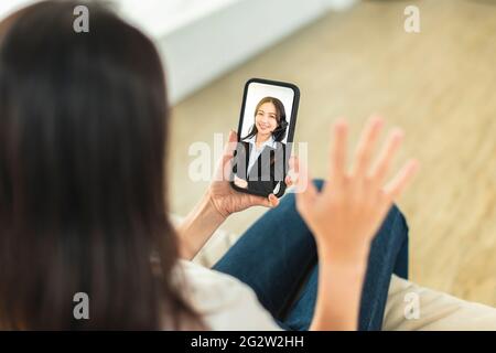 Giovane donna seduta sul divano con il telefono cellulare, videochiamata, discussione con i colleghi aziendali in telelavoro Foto Stock