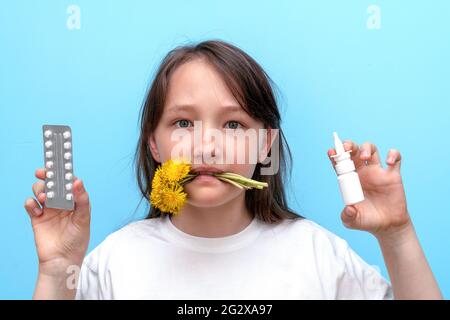 Ritratto di una bambina con fiori nei denti e pillole antiallergiche e spruzzare nelle mani su uno sfondo blu. Il concetto di polline stagionale Foto Stock